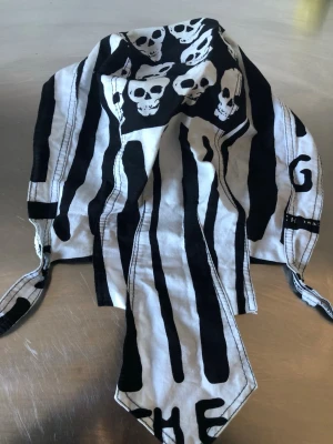 Svartvit dödskalle-bandana - Cool bandana i svartvitt med dödskallemönster och ränder. Tillverkad i tunn bomull, perfekt att knyta runt huvudet. Unik design med tryckta detaljer och text. Passar dig som vill sticka ut med din stil.