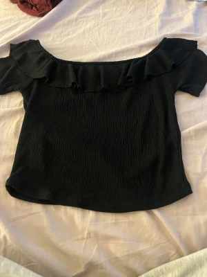 Svart offshoulder topp med volang - Trendig svart offshoulder topp från RA med volangdetalj längs urringningen. Toppen är ribbad och har korta ärmar, perfekt för en snygg och avslappnad look. Tillverkad i ett mjukt och stretchigt material som sitter bekvämt.