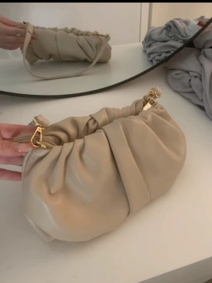 Beige axelremsväska i skinnimitation - Trendig beige handväska med mjuk, rynkad design och guldfärgade metalldetaljer.
