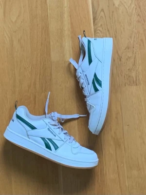 Vita Reebok sneakers med gröna detaljer - Snygga vita sneakers från Reebok med gröna detaljer på sidan och logga på hälen. Skorna har klassisk låg profil, vita snören och en slitstark sula. Perfekta för dig som gillar retrostil och vill ha något som funkar till det mesta.