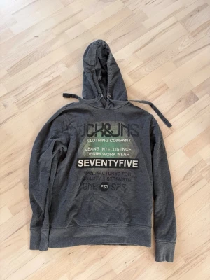 Grå hoodie från Jack & Jones - Snygg grå hoodie från Jack & Jones med tryck i grönt, vitt och svart på bröstet. Klassisk huva med dragsko och ribbade muddar vid ärmar och nederkant. Perfekt för en avslappnad streetwear-look. Materialet känns mjukt och bekvämt.