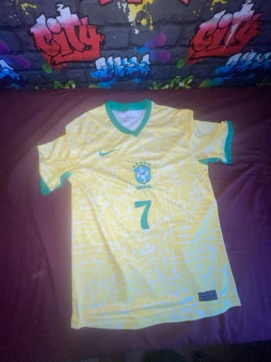 Brasilien matchtröja Vini Jr. #7 Nike - Gul Brasilien matchtröja från Nike med gröna detaljer, nummer 7 och Vini Jr. på ryggen. Tröjan har ett grafiskt mönster, rund halsringning och korta ärmar. Perfekt för dig som älskar fotboll och vill stötta Brasilien.