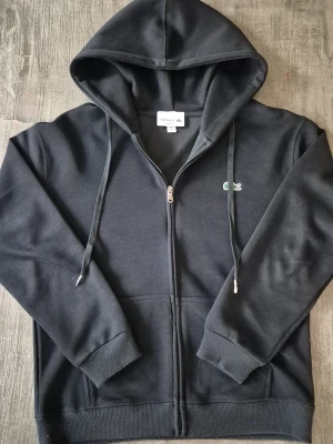 Svart zip hoodie från Lacoste - Snygg svart hoodie från Lacoste med dragkedja framtill och klassisk krokodil-logga på bröstet. Hoodien har huva med dragsko, ribbade muddar och fickor fram. Tillverkad i mjukt bomullsmaterial som känns skönt mot huden. Perfekt för en avslappnad och stilren look.