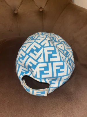Fendi blåvit keps med FF-mönster - Säljer en riktigt snygg keps från Fendi i blått och vitt med det klassiska FF-logomönstret över hela kepsen. Justerbar baktill för perfekt passform. Tillverkad i slitstarkt tyg som känns lyxigt och trendigt. Perfekt accessoar för dig som gillar exklusiv streetstyle.