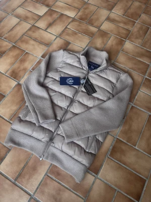 Beige cardiganjacka från Leon Marcel - Snygg beige cardiganjacka från Leon Marcelmed quiltad front och stickade ärmar samt rygg. Jackan har dragkedja framtill, ståkrage och ribbade muddar. Perfekt för lager på lager och en stilren look.