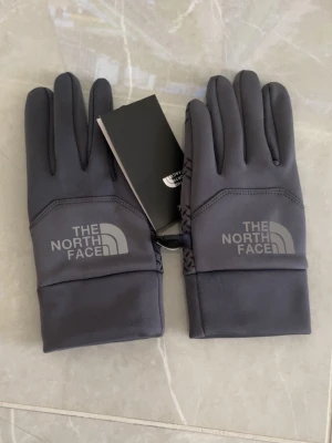 Grå sporthandskar The North Face - Grå sporthandskar från The North Face med logga på ovansidan och greppvänligt mönster i handflatan. Handskarna har touchfunktion på fingertopparna och breda muddar för extra komfort. Perfekta för utomhusaktiviteter under kalla dagar.