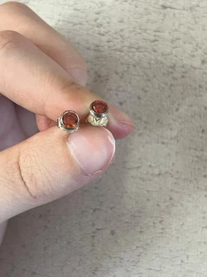 Silvriga örhängen med röd sten - Säljer ett par små silvriga örhängen med rund röd sten i mitten. Klassisk och enkel design som passar till många stilar. Stiften är i metall och stenen har en glansig finish som fångar ljuset snyggt.