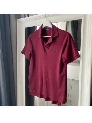 Vinröd pikétröja från Polo Ralph Lauren - Klassisk vinröd pikétröja från Polo Ralph Lauren med korta ärmar, krage och knappslå. Tröjan har den ikoniska broderade loggan på bröstet och är tillverkad i mjuk bomull. Perfekt för en avslappnad men stilren look.