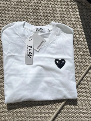 Vit Comme des Garçons PLAY t-shirt - Säljer en vit t-shirt från Comme des Garçons PLAY med det ikoniska svarta hjärtat med ögon broderat på bröstet. T-shirten har rund hals och korta ärmar. Tillverkad i mjuk bomull och har en clean, minimalistisk look.