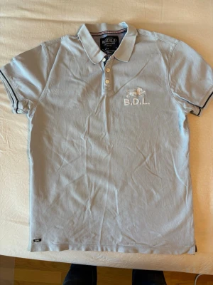 Ljusblå pikétröja från Bondfield - Snygg ljusblå pikétröja från Bondfield med vit brodyr och texten B.D.L. på bröstet. Klassisk krage med knappar och korta ärmar. Perfekt för en avslappnad men stilren look.
