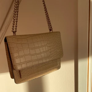 Axelremsväska - Säljer min beiga väska från Nelly då den inte längre kommer till användning. Den är rymlig och perfekt för både fest och vardag. Kjedjan har blivit lite missfärgad på vissa ställen men inget man tänker på, kolla bild två. Säljer för 120+ frakt