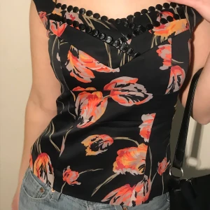 Svart topp med mönster, KAREN MILLEN - Figursydd topp med snygga detaljer vid bysten. Märket är Karen Millen och storleken 36 (UK-10, US-6). Plagget slutar precis strax under naveln på mig. Det finns en dragkedja på sidan av plagget. Färgen är svart med röda/orangea detaljer. Sparsamt använd i mycket bra skick💞!