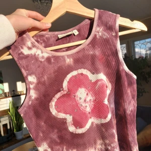 Crop top med blomma - Ett batikfärgat linne med en rosa blomma🌸🌸 blomman har även små paljetter på sig vilket är fint och den passar verkligen in i early 2000's viben! Står L men passar definitivt mindre också (jag är S)