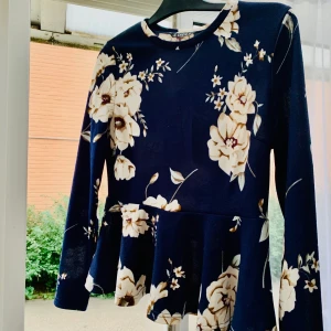 Blus - En supergullig blus som man kan använda till flera olika tillfällen. Den är marinblå med krämvita blommor och den har fina volanger nedtill. Blusen är i storlek L men passar även bra på folk som har S och M. Den är aldrig använd!