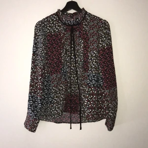  - Fin blus i blommigt mönster i rött blått svart och grönt. Fina veck sömmar längs mitten av blusens framsida. Snördetalj på framsidan som går att knyta. Knäppning på baksidan av blusen. Skönt tunt material i 100% polyester. Köpt på Bik Bok och endast använd 1 gång. Betalning sker via Swish och kunden står för frakten. 