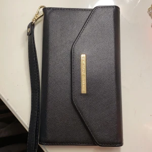 Mobilplånbok från Ideal of Sweden - Svart mobilplånbok från Ideal of Sweden för iPhone 8 i mycket fint skick. Magnetisk så mobilen lätt kan tas ut och sättas tillbaka. Gjord för iPhone 8 men funkar för andra mobiler också, bara det att kamerahålet inte kommer sitta rätt då. 70kr + frakt!