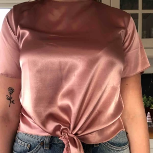  - Snygg t-shirt från Nelly i ett fint rosa blankt tyg. Med snygg knytning fram samt knapp bak i ryggen. Knappt använd så i fint skick. Köparen står för frakt🥰