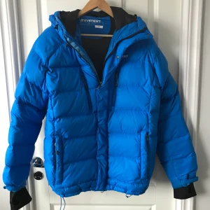 Everest jacka strl s-m - En ljusblå Everest dunjacka. Modellen har 2 fickor, justerbar midja, värmande muddar i ärmslut samt avtagbar luva. Jackan är vind och vattentät. Använd fåtal gånger därav i väldigt bra skick! Strl S men nassar jättebra som M också. Köparen står för frakten.