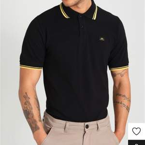 Svart Piké T-shirt med gula detaljer från Alpha Industries köpt 500kr från Zalando säljes för 150kr.