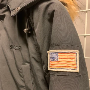 Svea jacka - Varm vinterjacka från svea! Ljuset på bilderna får den att se grå ut men den är svart. Säljer pga för liten. Dragkedjan är lite sönder längst ner men går ändå att stänga, har även knappar. 