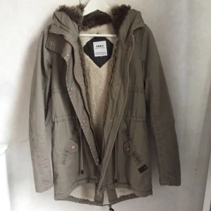 - Grå/grön vinterjacka med pälsimitation i luvan och insidan av jackan. Jackan har hög krage och detaljer i koppar/rosé. Använd mycket sparsamt, därför i väldigt fint skick! Betalning sker via swish! 
