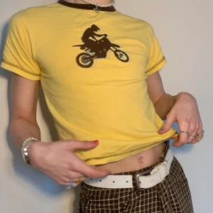 Gul y2k babytee med motorcykel tryck - As cool gul baby tee med brun contrast stitching längst ner, komplimenterande bruna kanter på ärmarna och kragen och ett brunt motorcykel tryck med ljusblå outlines ✨ så snygg till alla lågmidjade jeans och byxor eftersom färgerna är så neutrala!  