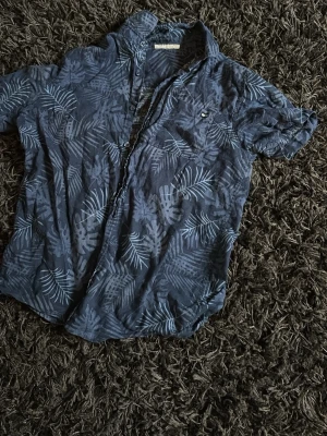 Blå skjorta med bladmönster - Säljer en blå skjorta med korta ärmar och ett snyggt bladmönster i ljusare blå toner. Skjortan har knappar framtill och en bröstficka. Perfekt för dig som vill ha en avslappnad men ändå stilren look.        Pris kan diskutaras behöver få den såld så fort som möjligt 