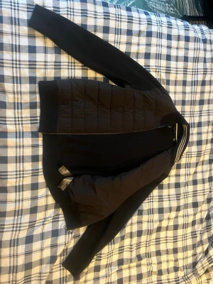 Svart cardiganjacka med quiltad front från Ted Baker - Snygg svart cardiganjacka med quiltad, vadderad front och stickade ärmar samt rygg från Ted Baker. Dragkedja framtill och ribbade muddar. Perfekt för lager-på-lager och ger en modern look till din outfit.