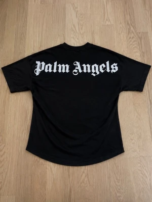 Svart Palm Angels t-shirt - Säljer en svart t-shirt från Palm Angels med stor vit logga på ryggen och mindre logga vid halsen fram. T-shirten har en loose passform och är tillverkad i mjuk bomull. Perfekt för dig som gillar streetwear och vill sticka ut.
