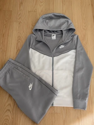 Grå och vit Nike loungewear-set - Snyggt loungewear-set från Nike i grått och vitt. Setet består av en långärmad hoodie med dragkedja och matchande byxor, båda med Nike-logga. Perfekt för chill dagar hemma eller när du vill vara bekväm men ändå se fräsch ut.