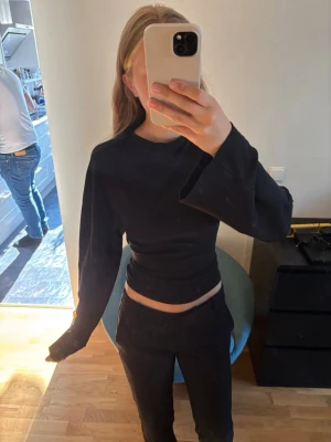 Svart omlottopp med knyt och vida ärmar - Säljer en svart omlottopp med långa, vida ärmar och knyt i ryggen. Toppen är croppad och har en tight passform som framhäver midjan. Storleken passar alla då man väljer hur den ska sitta med knytningen i ryggen. Använd 3 ggr men inga skador alls.