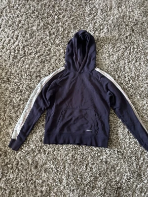 Mörkblå hoodie med vita ränder GANT - Snygg mörkblå hoodie från GANT med huva och magficka. Vita ränder med GANT-logga längs båda ärmarna ger en sportig vibe. Perfekt för en avslappnad stil. Tillverkad i mjukt bomullsmaterial.