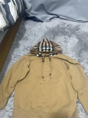 Byrberry hoodie, pris går att diskutera  - Säljer en beige hoodie med stor ficka framtill och en huva med klassiskt rutmönster i brunt, svart, vitt och rött. Hoodien har dragsko med bruna detaljer och är gjord i mjukt material. Perfekt för en chill och stilren look.