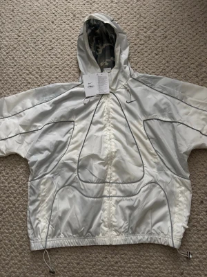 Nike X Corteiz vit reflekterande vindjacka - Snygg vit vindjacka från Nike med reflekterande detaljer som lyser upp i mörker. Jackan har huva med kamouflagemönstrat foder, dragsko i nederkant och en sportig design med svarta sömmar. Perfekt för dig som vill sticka ut och synas på kvällen.