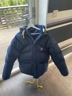 Fjällräven jacka marinblå - Säljer en marinblå dunjacka från Fjällräven med huva och gula dragskor. Jackan har två stora fickor framtill, Klassisk Fjällräven-logga på bröstet. Perfekt för kalla vinterdagar. Den är lite stor i storlek. Priset kan diskuteras