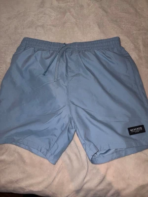 Ljusblå badbyxor från McKenzie - Snygga ljusblå badbyxor från McKenzie med elastisk midja och dragsko för skön passform. Klassisk kort modell med diskret logga på vänster ben. Perfekta för stranden eller poolen. Tillverkade i snabbtorkande material.