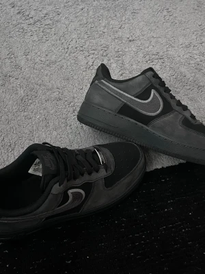 Svart/gråa Nike Air Force 1 sneakers - Snygga svarta Nike Air Force 1 sneakers med grå swoosh och detaljer. Skorna har klassisk låg siluett, svarta skosnören och ovandel i mocka och läder. Perfekta för dig som gillar stilrena och tidlösa sneakers med streetkänsla.