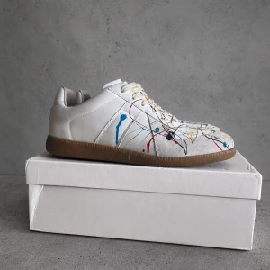 Maison margela - Unika vita sneakers med färgstänk i blått, rött, gult och svart. Skorna har en klassisk låg siluett och en brun gummisula. Tillverkade i slätt läder med vita snören och diskreta paneldetaljer. Perfekta för dig som vill sticka ut med din stil. Använda en gång 