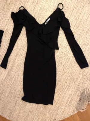 Svart långärmad klänning med volang - Snygg svart klänning med långa ärmar och offshoulder-design. Klänningen har volangdetaljer längs ringningen och axlarna, vilket ger en trendig och feminin look. Perfekt för dig som gillar stilrena och eleganta plagg.