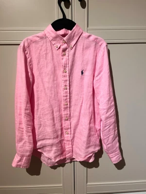 Ralph lauren - Rosa linneskjorta  - Snygg rosa skjorta i linne med klassisk krage och knappar framtill. Skjortan har lång ärm och en broderad logga på bröstet. Perfekt för en fräsch och stilren look.