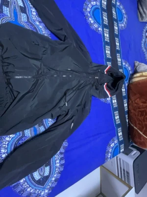 Moncler - Snygg svart vindjacka med huva och dragkedja framtill. Jackan har en diskret logga på ärmen och en randig detalj i rött, vitt och blått på huvan. Perfekt för blåsiga dagar och enkel att matcha med olika outfits.