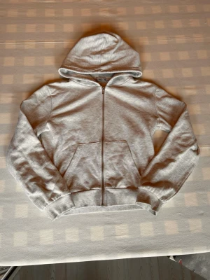 Ljusgrå zip hoodie med huva - En stilren ljusgrå hoodie med dragkedja framtill och stor ficka på magen. Hoodien har ribbade muddar vid ärmslut och nederkant samt en klassisk huva. Perfekt för en avslappnad och bekväm look.