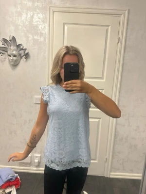 Ljusblå spetsblus med volangärm - Supersöt ljusblå blus i spets med volangdetaljer vid ärmarna. Blusen är ärmlös och har en rak passform med fina spetsmönster över hela plagget. Perfekt att styla med jeans eller kjol för en fräsch look.