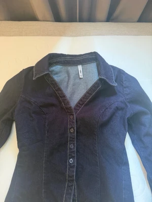 Mörkblå figursydd jeansjacka - Snygg mörkblå jeansjacka med figurnära passform och klassisk krage. Jackan har långa ärmar, knäppning framtill med fem knappar och kontrastsömmar som ger en cool look. Perfekt för dig som vill ha en stilren och trendig jeansjacka.