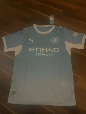 Manchester City matchtröja Puma - Ljusblå Manchester City matchtröja från Puma med vita detaljer och klubbmärke på bröstet. Tröjan har Etihad Airways-tryck på framsidan och är tillverkad i ett lätt och ventilerande material. Perfekt för fotbollsträning eller att bära på matchdag.