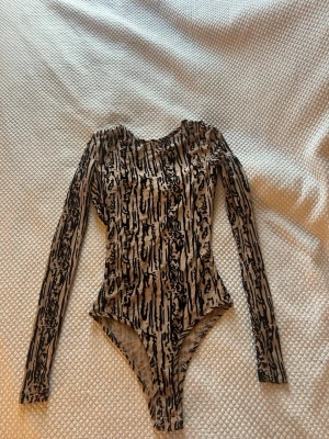 Långarmad mönstrad body  - Snygg body med lång ärm i beige och svart meshmaterial. Unikt abstrakt mönster och stor öppen rygg som ger en cool look. Tight passform och rund halsringning. Perfekt för dig som vill sticka ut lite extra. Helt oanvänd 