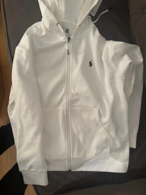 Vit hoodie med dragkedja från Polo Ralph Lauren - Snygg vit hoodie med dragkedja från Polo Ralph Lauren. Klassisk design med huva, fickor framtill och den ikoniska mörkblå loggan broderad på bröstet. Tillverkad i mjukt bomullsmaterial och har ribbade muddar vid ärmslut och nederkant.