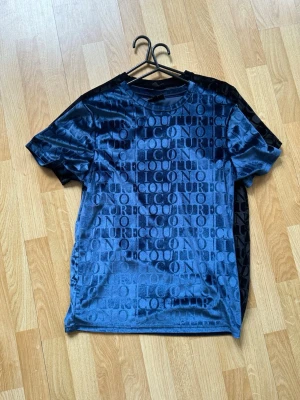 2 för 1 - 2 för 1. Två t-shirts i sammet med glansig finish, en i mörkblå och en i svart. Båda har ett upprepat textmönster med ordet 'ICON' och 'COUTURE' över hela tröjan. Klassisk rund halsringning och korta ärmar. Perfekt för dig som gillar statement-plagg.