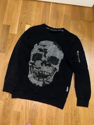 Svart Philipp Plein tröja med döskalle - Cool svart sweatshirt från Philipp Plein med stor glittrig döskalle framtill. Tröjan har rund halsringning, långa ärmar och en liten ficka med dragkedja på ena ärmen. Perfekt för dig som gillar edgy och unik stil.