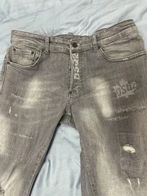 Grå slitna jeans med tryck - Säljer ett par grå jeans med slitningar och tryckdetaljer på framsidan. Jeansen har fem fickor, bälteshällor och dragkedja. Materialet är klassisk denim och de har en cool, tvättad look med vita detaljer och tryck på ena benet.